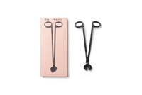 product shot of exterior pink box with black text, boy smells logo in black and black image of wick trimmer , next to box is metallic black wick trimmer with engraved boy smells logo against a neutral background. 